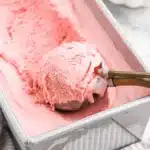 A large tray of creamy Raspberry Ice Cream Recipe with a metal scooper lifting a perfect scoop.