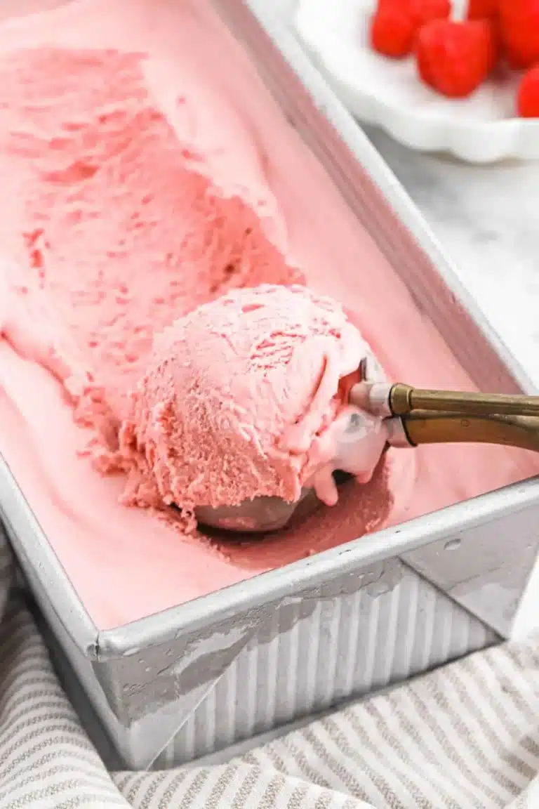 A large tray of creamy Raspberry Ice Cream Recipe with a metal scooper lifting a perfect scoop.