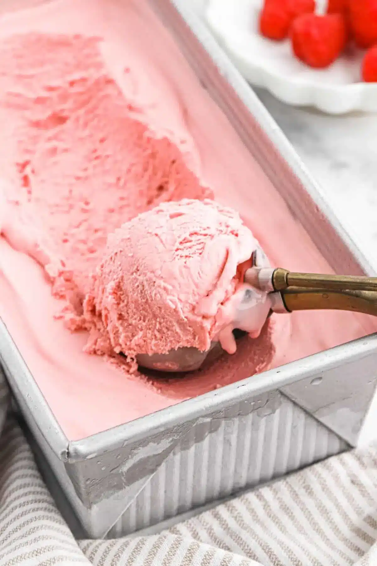 A large tray of creamy Raspberry Ice Cream Recipe with a metal scooper lifting a perfect scoop.