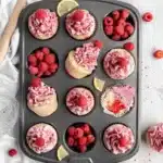 Freshly baked Raspberry Lemon Poke Cake Cupcakes Recipe treats arranged in a muffin tin with whole raspberries nestled in empty slots against a clean white backdrop.