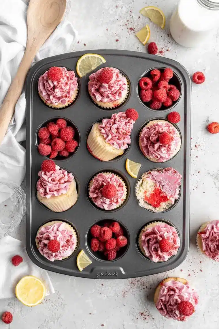 Freshly baked Raspberry Lemon Poke Cake Cupcakes Recipe treats arranged in a muffin tin with whole raspberries nestled in empty slots against a clean white backdrop.