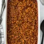 Large dish of homemade baked beans in a white casserole dish with a black spoon on the side