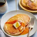 Maple syrup being poured from a pitcher over a plate of pancakes